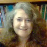 Headshot of Kelly Ace in front of a bookshelf. She has wavy brown and gray hair and is smiling.