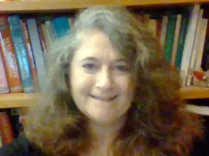 Headshot of Kelly Ace in front of a bookshelf. She has wavy brown and gray hair and is smiling.